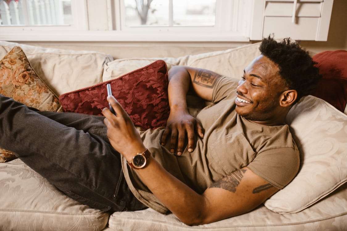 A black man is lying on a sofa, looking at the phone with a smile
